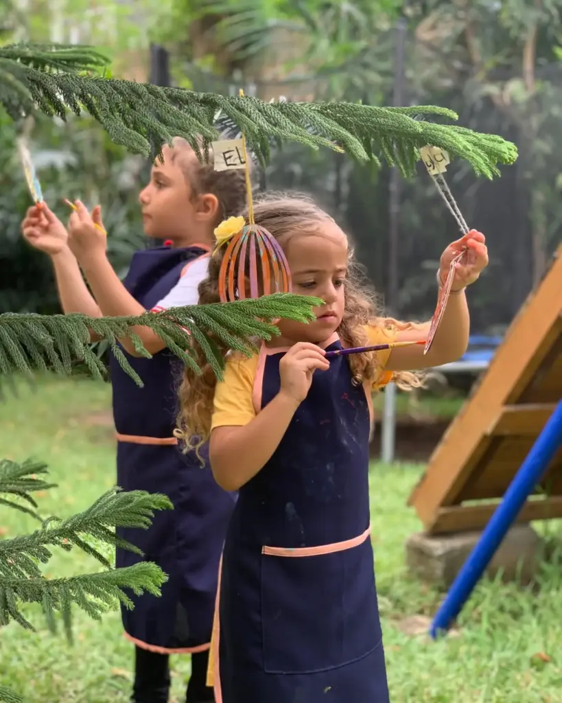 Two girls in aprons painting egg ornaments hanging from a garden pine tree